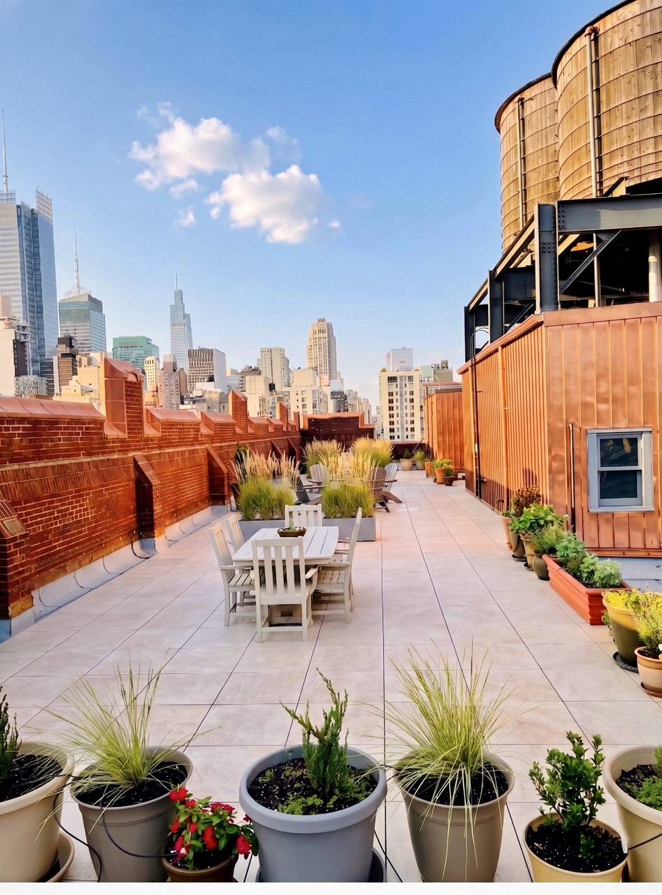 The Glass Farmhouse rooftop, looking east toward the Empire State Building, with planters, a wooden water tower at right, and tile pavers running the length of the deck